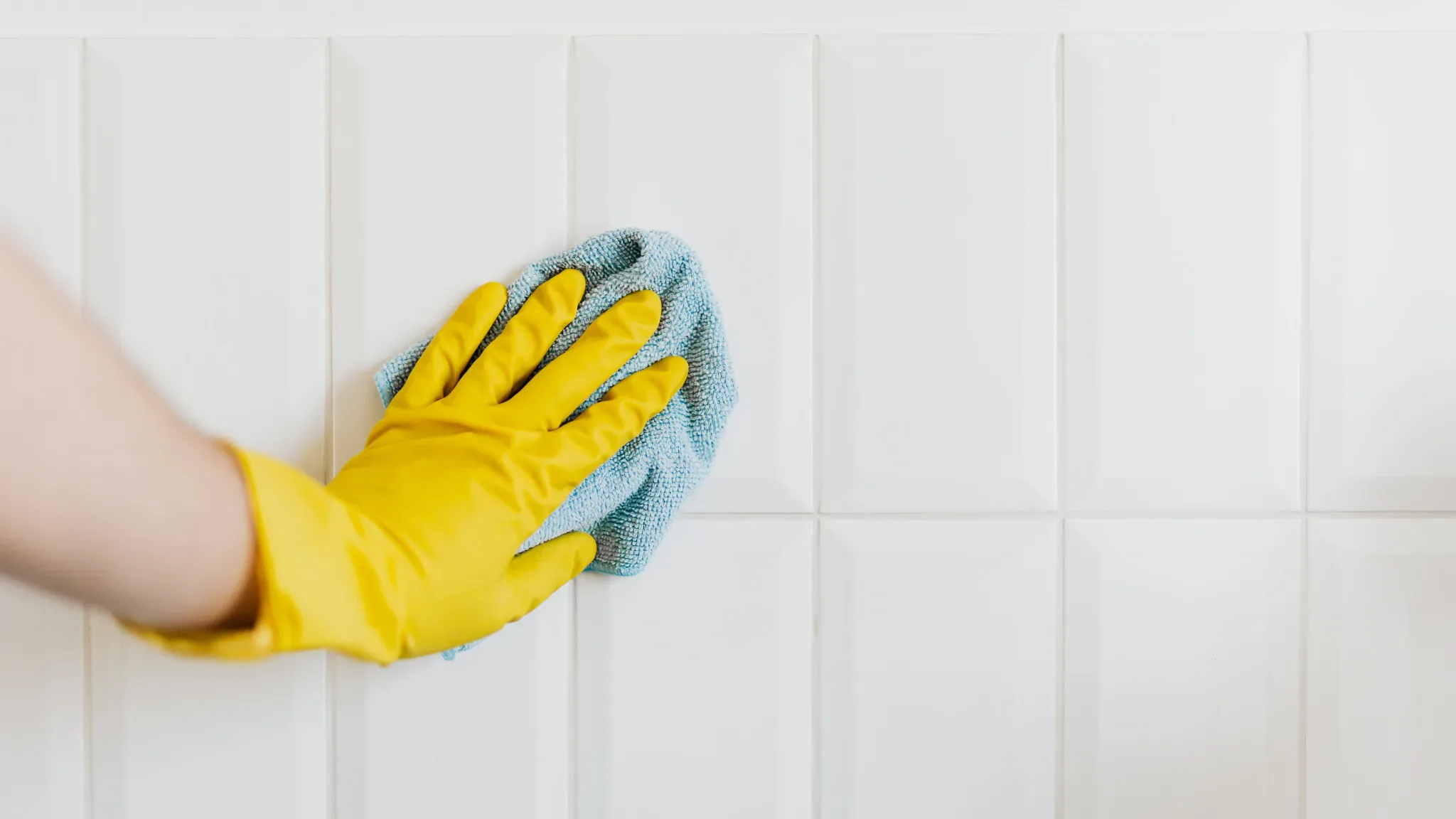 A person wearing yellow rubber gloves cleans white tile wall with a blue microfiber cloth, representing professional house cl