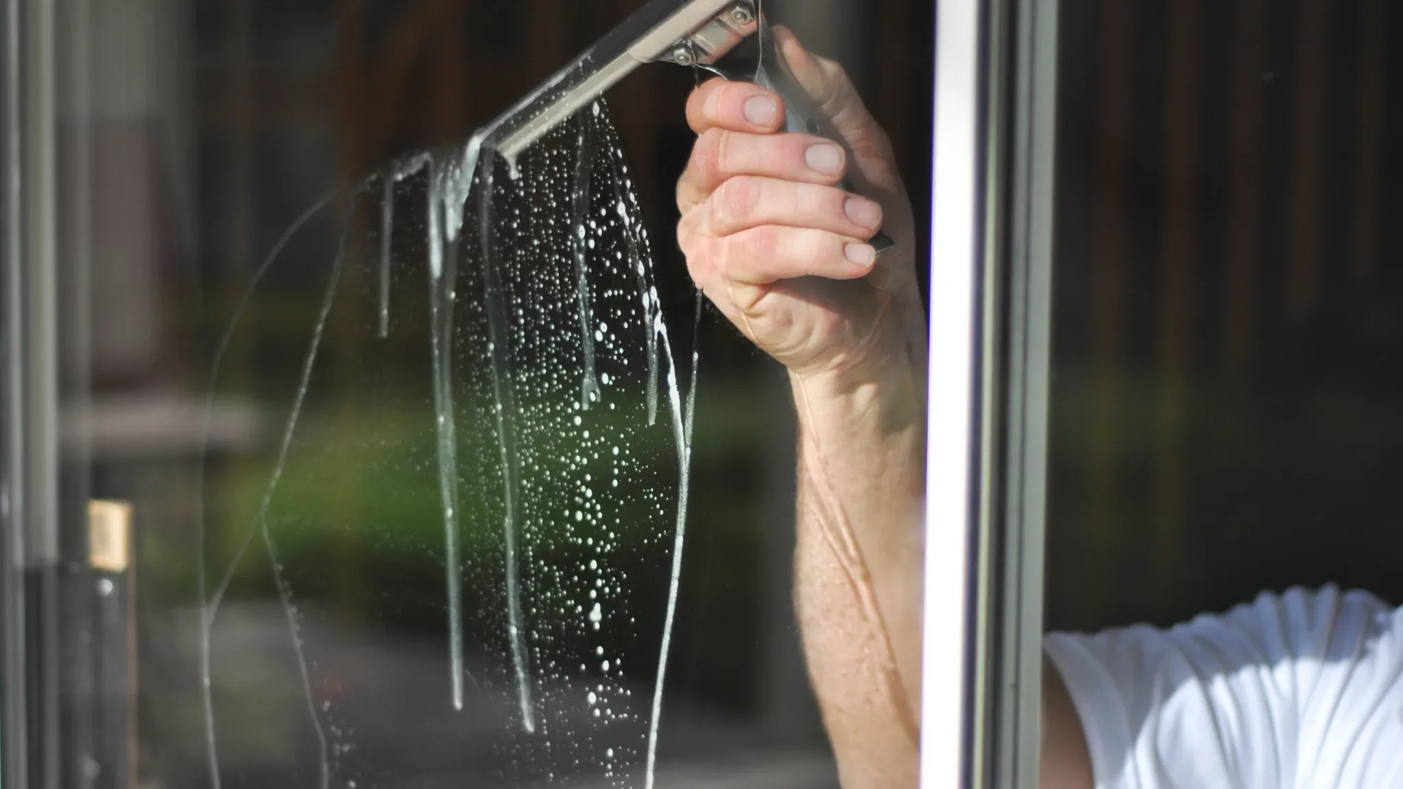 Professional window cleaner using squeegee to wash glass surface with water spray, demonstrating residential cleaning service