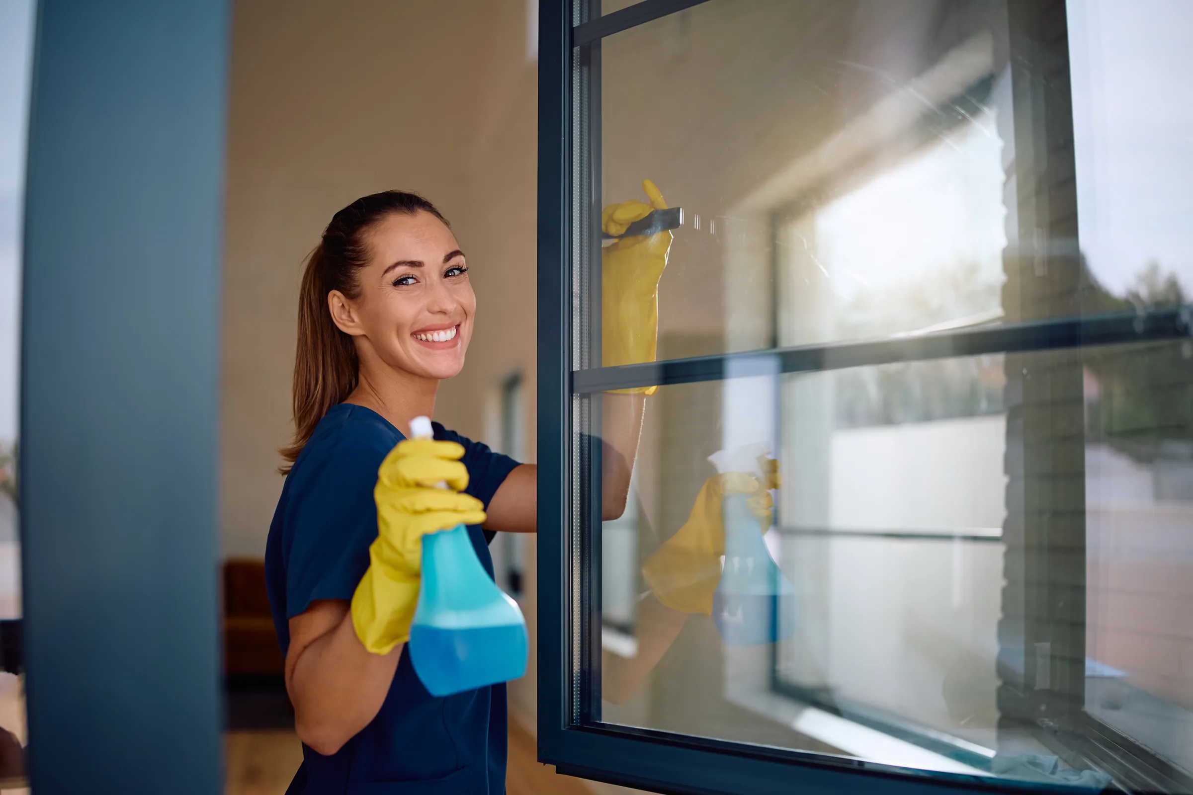 Smiling woman in yellow gloves spraying window cleaner on glass window.